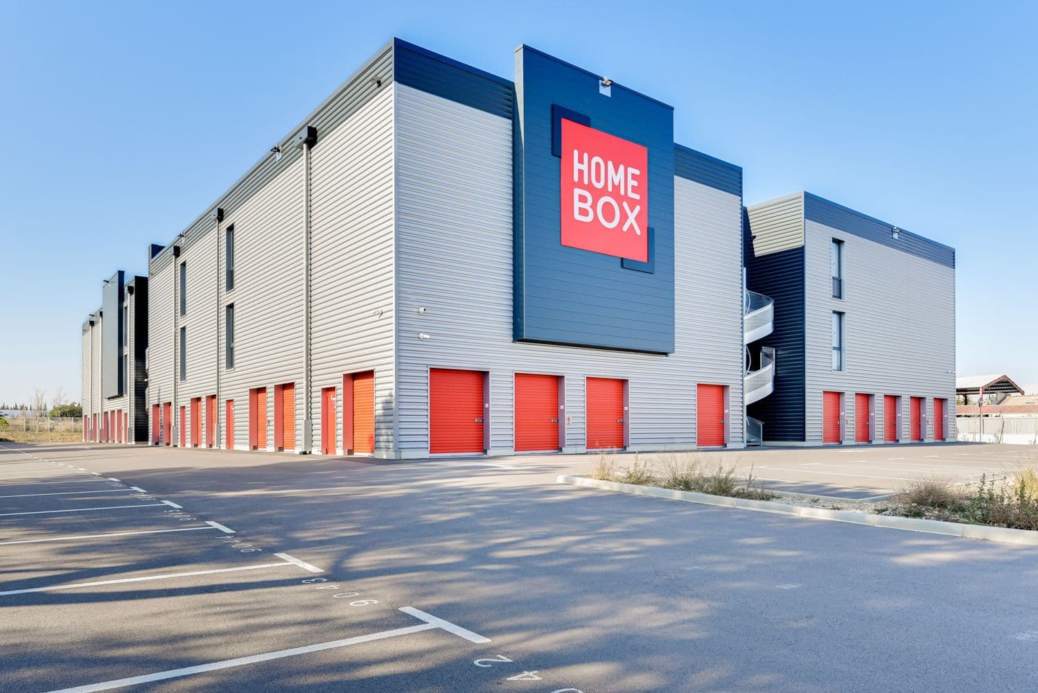 Vue extérieure d’un centre HOMEBOX moderne avec façade grise et portes de box rouges, disposant d’un grand parking.